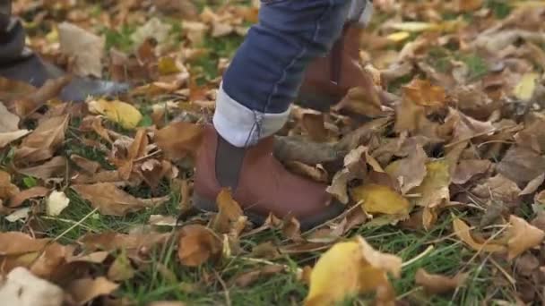 Close-up Een kleine jongen van een jaar oud, met zijn moeder, in bruine laarzen en blauwe jeans, leert wandelen in een herfstpark bedekt met geel, goud en rood gebladerte — Stockvideo