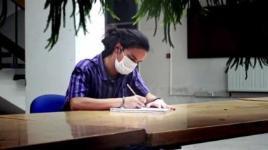 Uzun saçlı, soyutlanmış ve maskeli bir üniversite öğrencisi kütüphanede bir ağacın önünde tek başına notlar alıyor.