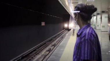 İzole edilmiş, uzun saçlı, yüz kalkanı ve maskesi takan zeki genç adam metronun yalnız gelmesini bekliyor.