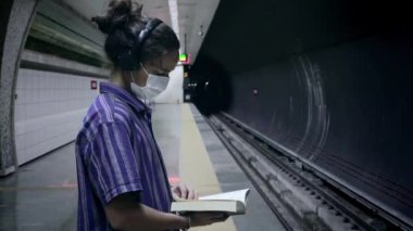 Maskeli, uzun saçlı yetişkin yürüyüşleri, kulaklığından müzik dinlemek ve metronun gelmesini beklerken edebiyat kitabı okumak.