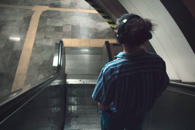 Metroda yürüyen merdivende duran ve kulaklıkla müzik dinleyen uzun saçlı genç adam.