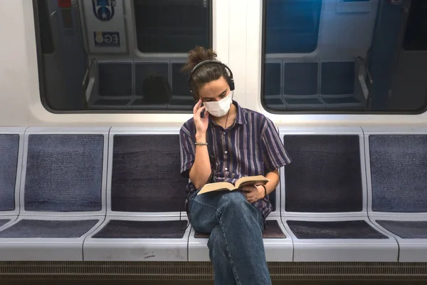 Isolated and masked smart looking young adult man listening to music and reading a literature book while traveling in the subway
