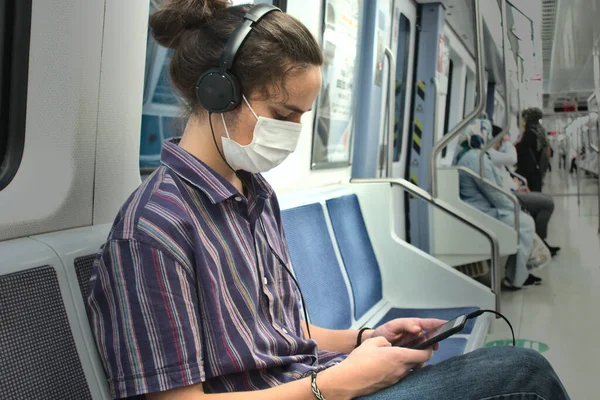 Uzun saçlı genç yetişkin milenyumu metroda telefonuyla oynarken kulaklığından müzik dinliyor sosyal mesafeli insanlarla seyahat ediyor.