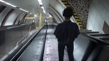 Metroda yürüyen merdivende duran ve kulaklıkla müzik dinleyen uzun saçlı genç adam.
