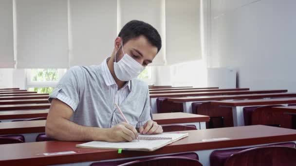 一个年轻的黑发男子独自一个人在大学的教室里 在社交场合独自学习 戴着外科口罩 写东西给他的书 教室里空无一人 这是由于考罗那病毒的流行 — 图库视频影像