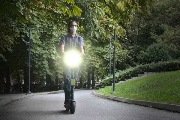 Uzun saçlı, genç, maskeli, trende seyahat eden ve gece vakti parkta farları açık, çevre dostu elektrikli scooterlar giyen bir yetişkin.