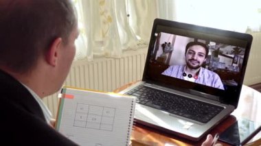 Evde çalışan orta yaşlı bir adam bilgisayarını genç bir meslektaşıyla kullandığı bir video görüşmesinde not alıyor.
