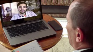 Evde çalışan gülümseyen orta yaşlı bir adam laptopunu genç meslektaşıyla birlikte kullandığı bir video görüşmesini not alıyor.
