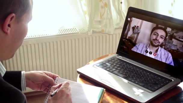 若い同僚とノートパソコンを使っているというビデオ通話でメモを取りながら家で働いているシニア大人の男 — ストック動画