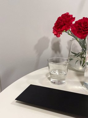 Kırmızı şakayıklar, yakınlarda siyah menü bulunan beyaz bir masadaki cam vazoda sergileniyor..