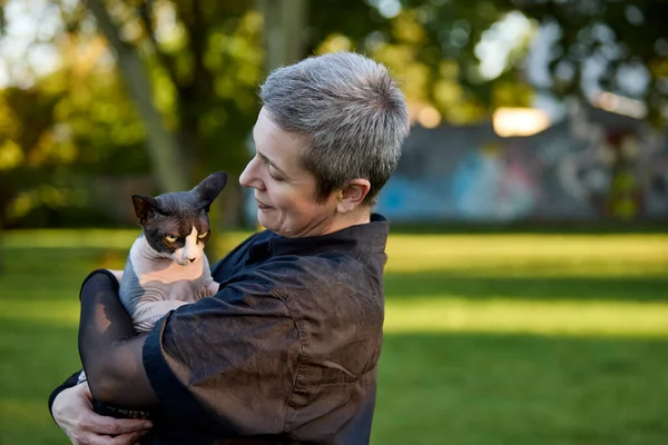 Kısa gri saçlı neşeli bir kadın Kanadalı Sphynx kedisini parkta sallıyor. Görüntü neşe, şefkat ve evcil hayvan ve sahibi arasındaki anlamlı arkadaşlığı gösteriyor..