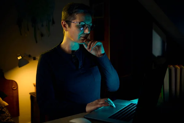 Orta yaşlı bir kadın elinde telefonuyla uzaktan kumandayla karanlık bir ofiste çalışıyor. Görüntü, ev kültürü ve gece geç saatte yapılan iletişimi temsil ediyor..