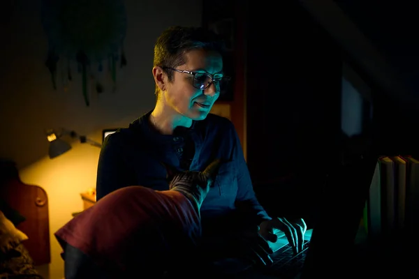 Orta yaşlı bir kadın gece geç saatlerde evinde Kanadalı Sphynx kedisi kucağında dinlenirken çalışıyor. Loş ofis ışıklandırması evde uzaktan çalışma ve arkadaşlık karışımını vurguluyor.