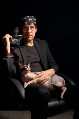 Siyah elbiseli bir fotoğrafçının yaratıcı portresi kucağında Sphynx kedisiyle karanlık bir stüdyoda kamera tutuyor. Görüntü sanatsal yaşam tarzını ve evcil hayvanlar arasındaki bağı yansıtıyor..