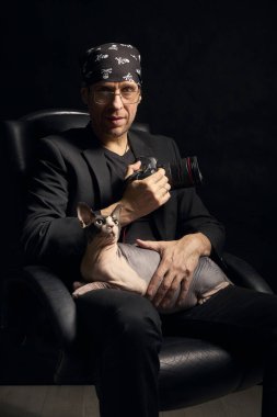 Sandalyede otururken Kanadalı Sphynx kedisini tutan bir adamın kapalı bir portresi. Siyah arkaplan ve dramatik ışıklandırma bağlantıyı, yaratıcılığı ve yaşam tarzı kimliğini vurguluyor.