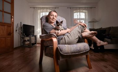 Orta yaşlı, kısa gri saçlı bir kadın, Kanadalı Sphynx kedisini sıcak bir oturma odasında elinde tutan tahta bir sandalyeye oturur. Pencereden gün ışığıyla aydınlatılır..