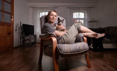 Orta yaşlı, kısa gri saçlı bir kadın Kanadalı Sphynx kedisiyle oturma odasında oturur ve sıcak bir arkadaşlık ortamı ve günlük ev hayatı yaratır..