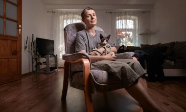 Gri saçlı orta yaşlı bir kadın ve Kanadalı Sphynx kedisi, arka plandaki perdelerden süzülen güneş ışığıyla, parlak bir oturma odasındaki sandalyeye sakince oturur..