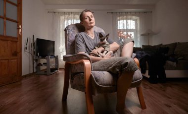 Gri kısa saçlı orta yaşlı bir kadın modern bir oturma odasında kucağında Kanadalı Sphynx kedisiyle rahatça oturur. Konforu, huzuru ve günlük arkadaşlığı somutlaştırır..