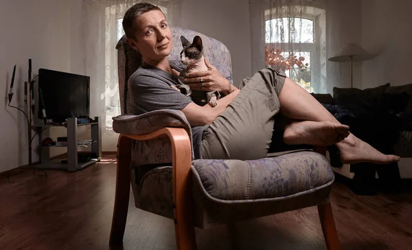 Orta yaşlı, kısa gri saçlı bir kadın kameraya bakarken Kanadalı Sphynx kedisini tutuyor, modern bir oturma odasında, ahşap zemin ve doğal ışıkla oturuyor..