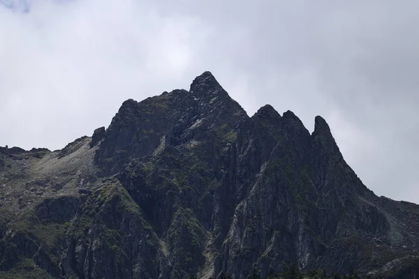 Dramatik bir şekilde yükselen, karanlık, sivri dağ zirveleri bulutlu bir gökyüzünü delip dokunulmamış doğanın saf güzelliğini ve gücünü vurgulayan heybetli bir manzara oluşturuyor.