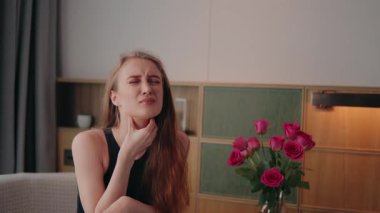 Sağlığı kötü olan genç bir kadın boğaz ağrısı çekiyor ve yutkunmaya çabalıyor, evde oturuyor ve ağır çekimde rahatsızlıktan yüzünü buruştururken nazikçe boynunu tutuyor.