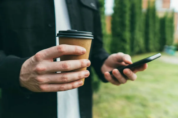 Adam akıllı telefonuyla mesaj yazıyor ve parkta sıcak kahve içiyor.