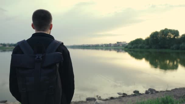 Hombre viajero con mochila de pie mirando el hermoso atardecer cerca del río — Vídeo de stock