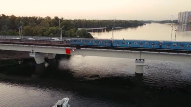 Tekne, gün batımında şehir bölgelerinde metro işleten bir köprünün altından geçiyor..