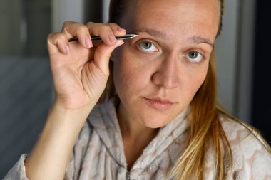 Sarışın bir kız banyoda cımbızla kaşlarını yoluyor. Odaklanma ve özen gösteriyor. Güzellik rutinleri, kişisel bakım ya da kişisel anları olan görsel yaşam tarzı için idealdir..