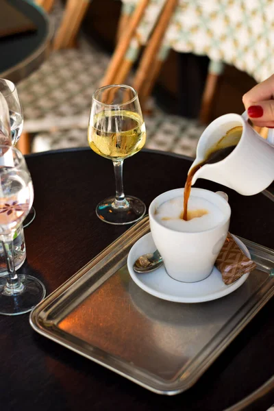 Bir kadının elini dikey çekimde süt köpüğüne kahve dökerken, masada beyaz şarap, örülmüş sandalyelerin Paris titreşimleri. Seyahat görüntüleri, yemek sahneleri ya da rahat bir yaşam tarzı için ideal..