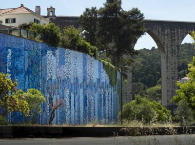 Bright blue graffiti mural on a high wall in the suburbs of Lisbon, Portugal near the road. Sunny weather and warm spring, behind the wall you can see an old stone bridge. Nature and a landscape.