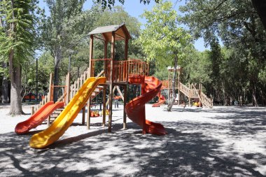 Vertical view of a wooden playground set with bright orange and yellow slides under trees. Useful for childhood, outdoor activity, park equipment, family recreation, safety design, and community themes.