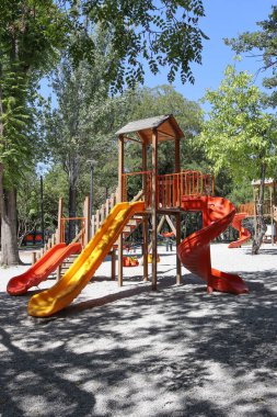 Vertical view of a wooden playground set with bright orange and yellow slides under trees. Useful for childhood, outdoor activity, park equipment, family recreation, safety design, and community themes.