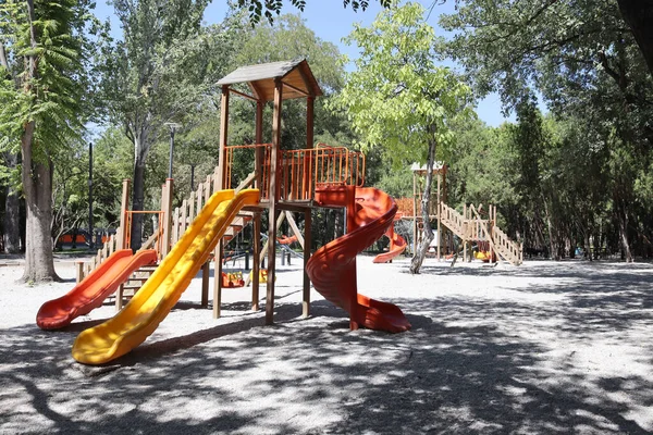 Vertical view of a wooden playground set with bright orange and yellow slides under trees. Useful for childhood, outdoor activity, park equipment, family recreation, safety design, and community themes.