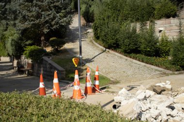 Orange cones and tape close a park path near landscaping and a cobblestone slope. Useful for detour, safety, access control, maintenance, and public space management themes.
