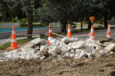 Orange traffic cones, warning tape and broken concrete marking a repair zone in a park walkway. Great for maintenance, hazard, safety, urban infrastructure and construction themes.