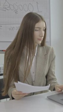 In a modern office, a young businesswoman diligently reviews important papers while working on her laptop