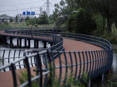 Curved pedestrian bridge with metal railings crossing over water with highway infrastructure in background