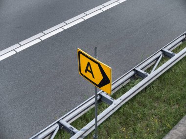 A harfli sarı yol işareti ve karayolu bariyerlerinin yanına monte edilmiş yön oku.
