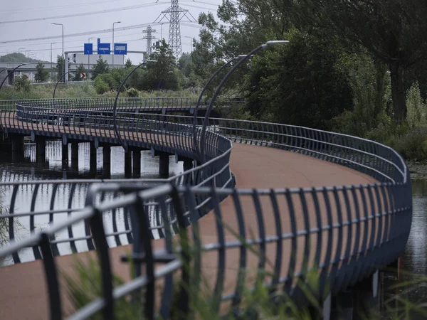 Curved pedestrian bridge with metal railings crossing over water with highway infrastructure in background