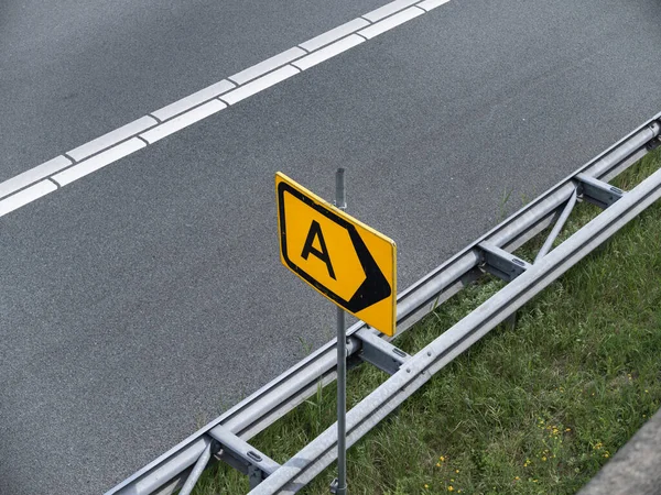 A harfli sarı yol işareti ve karayolu bariyerlerinin yanına monte edilmiş yön oku.