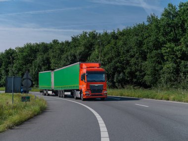 Orange truck with green trailer navigating curved road section with grass verges and trees