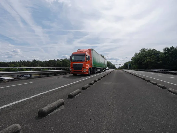 Orange truck with green trailer on highway with concrete barriers and overcast sky