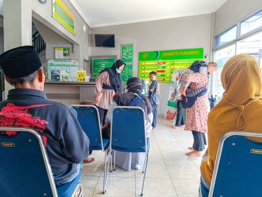 Kamu hizmeti tesisinde çok sayıda insan sırasını bekliyor, bazıları maske takıyor ve diğerleri sağlık hizmetleri veya kamu hizmeti içeriğine uygun personelle etkileşimde bulunuyor.
