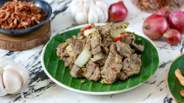 Yeşil muz yaprağı tabağında soğan ve biberli leziz biftek kızartması sunuluyor. Aperatif bir yemek sunumu yaratıyor. Yemek blogu ya da yemek tarifi postası için mükemmel..