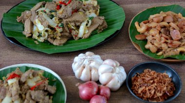 Ahşap bir yüzeye yerleştirilmiş kızarmış et yemekleri, sebzeler ve sarımsak soğanları, aşçılık içeriği ve yemekle ilgili projeler sunuyor..