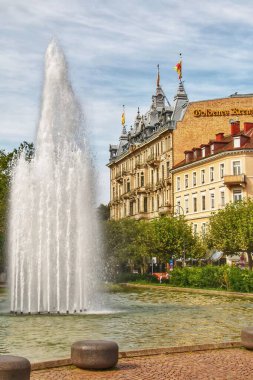 Görkemli bir çeşme, parçalı bulutlu bir gökyüzünün altındaki tarihi binalar tarafından çerçevelenmiş Baden-Badens merkez meydanının kalbine uzun bir su püskürtür. Kültür, mimari ve şehir zarafetinin canlı bir karışımı. Tarih: 3 Eylül 2023 