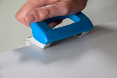 Mavi ve beyaz bir kağıt kesiciyi kavrayan, düz bir yüzeydeki beyaz bir çarşafı budayan bir elin yakın çekimi. Resim, ofis, okul veya zanaat ortamlarında tipik bir manuel kesim görevini göstermektedir..