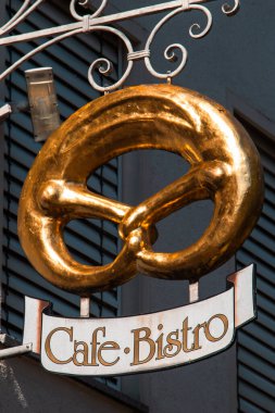 Decorative Cafe Bistro sign with a golden pretzel and stylized text, mounted on a wrought iron bracket outside a building with shutters. A visual symbol of traditional European cafe culture and baked goods. Date: 15 Sept. 2025